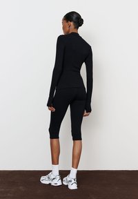 Fekete, hosszú ujjú sportfelső, hüvelykujjlyukakkal, fekete cropped leggings-szel párosítva. Fehér bokazoknival és szürke futócipővel viselve.