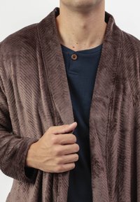 Hombre que lleva una camisa azul oscuro con un botón y un bata marrón texturizada, sujetando la bata cerrada con su mano derecha.