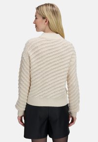 Frau mit langen blonden Haaren, die einen cremefarbenen strukturierten Strickpullover und schwarze Shorts trägt, mit dem Rücken zu einem neutralen Hintergrund.