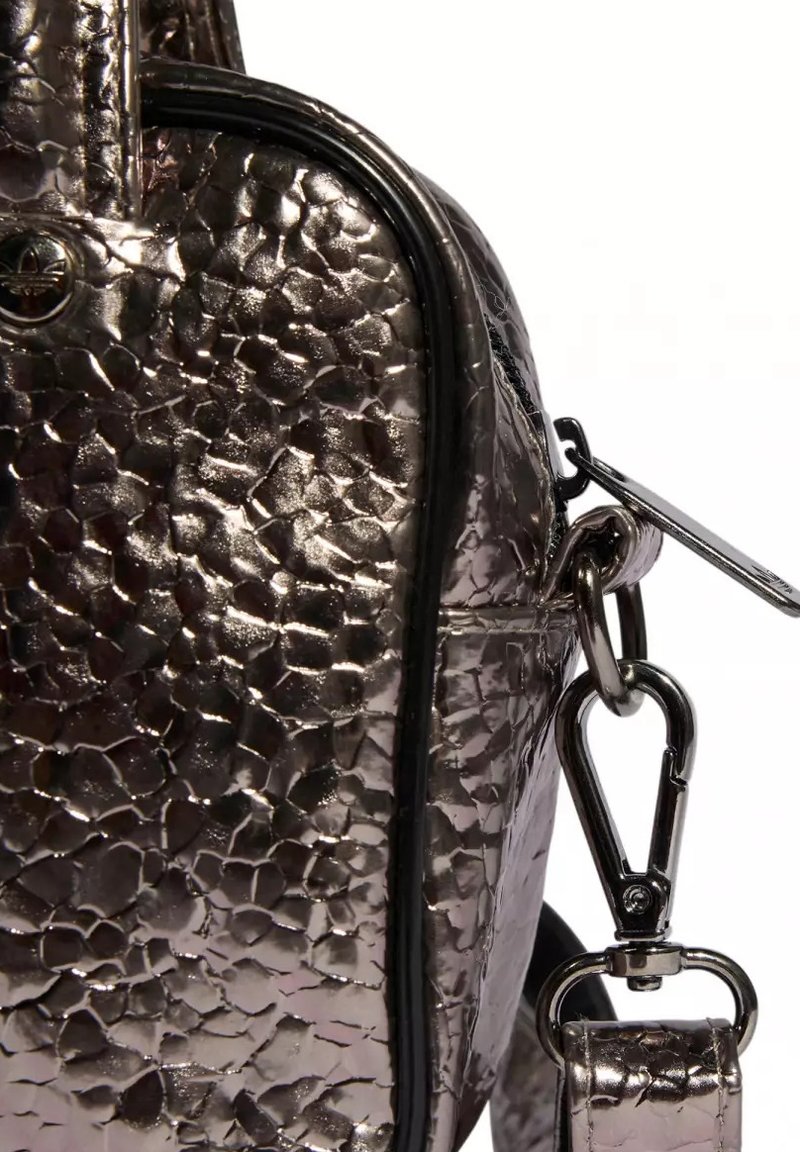 Primo piano dell’angolo di una borsa metallizzata con cerniera, rifinitura nera e chiusura girevole argentata che fissa la tracolla.