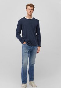 Dunkelblaues Langarmshirt mit Rundhalsausschnitt, kombiniert mit hellblauen Jeans. Modell trägt weiße Sneakers und steht vor einem weißen Hintergrund.