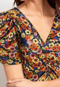 Un blouse floral avec une base marine et des fleurs colorées en rouge, jaune et violet. Design en V et détails froncés au centre.
