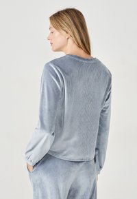 Sudadera de terciopelo azul con cuello redondo y mangas largas, que presenta una textura de canalé y un corte holgado, mostrada desde la parte posterior.