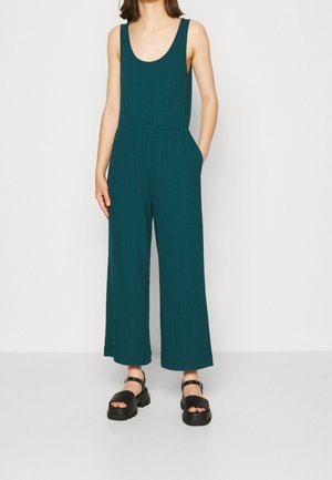 Woman wearing sleeveless dark green wide-leg jumpsuit with pockets and black chunky platform sandals standing against a plain background.