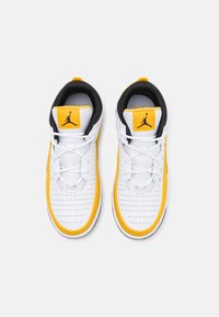 Vita och gula sportskor med svart foder, perforerat tåområde och Jumpman-logga på den gula tunga delen, sett uppifrån.