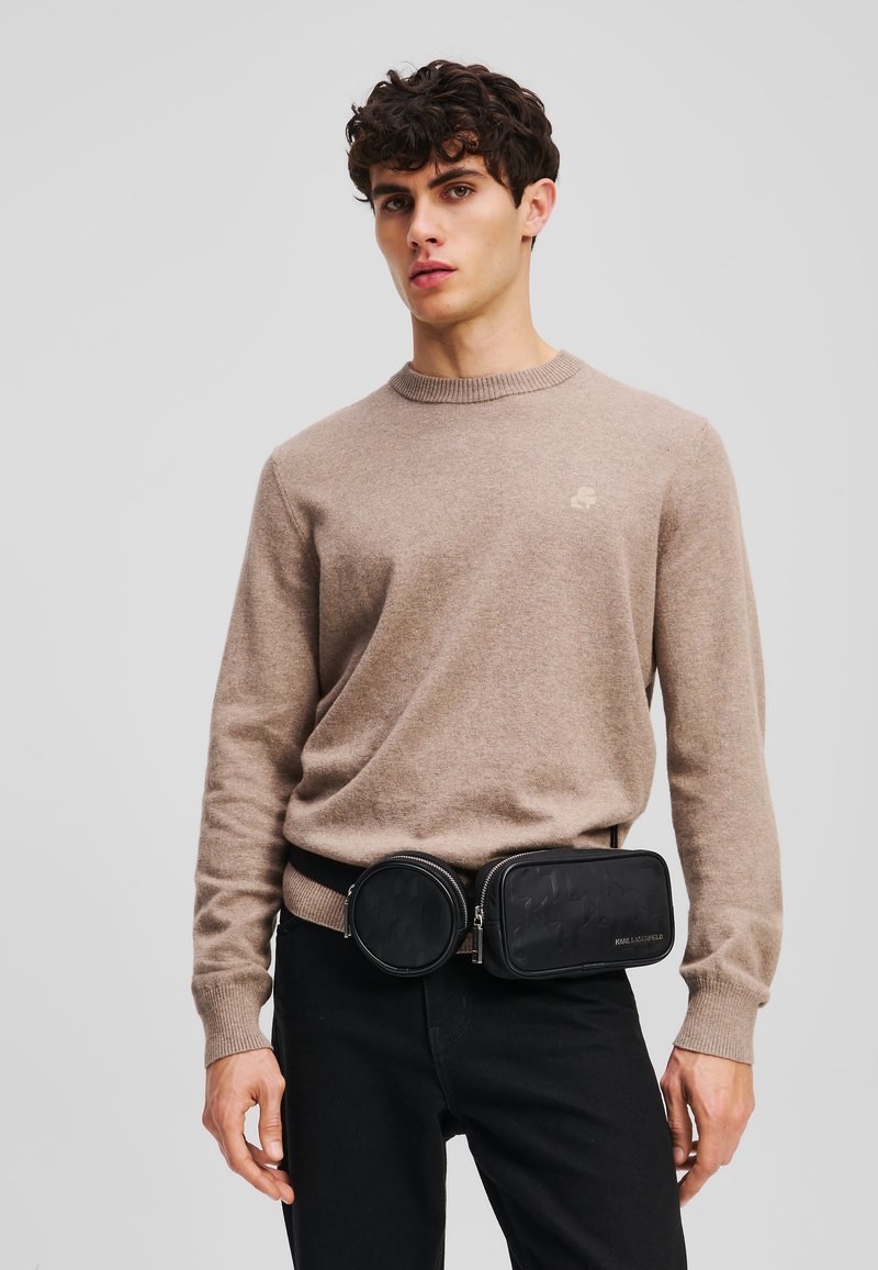 Maglione di lana marrone chiaro con scollo rotondo, abbinato a una cintura a tracolla nera con due scomparti, caratterizzata da un motivo strutturato e accessori in metallo argentato.