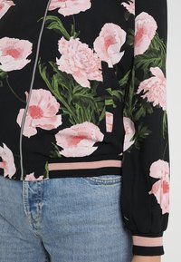 Giubbotto bomber nero con grande stampa floreale rosa, polsini e orlo a costine rosa. Apertura con zip, realizzato in tessuto morbido. Presenta maniche arricciate.