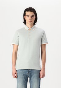 Polo en tissu vert clair avec un col et des poignets rayés orange et blanc, avec une patte de boutonnage à trois boutons et un petit logo.