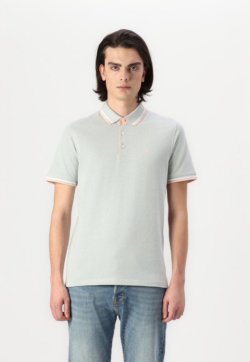 Polo en tissu vert clair avec un col et des poignets rayés orange et blanc, avec une patte de boutonnage à trois boutons et un petit logo.