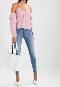 Punavalkoraidallinen olkapäät paljastava pusero, jossa on röyhelöhihat, yhdistetty vaalean sinisiin kapeneviin farkkuihin ja valkoisiin korkeakorkoisiin kenkiin, kädessä valkoinen laukku.