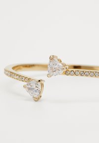 Bracciale rigido dorato con due pietre trasparenti a forma di cuore e una fila di piccoli cristalli su un lato; finitura liscia.