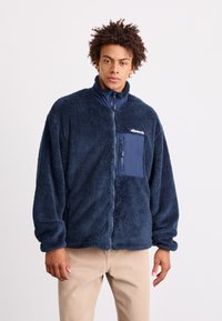 Mornarska flis jakna s visokim ovratnikom, zatvaranjem na zip i prednjim džepom na grudima. Ima meku teksturu i elastične rukave. Logo je na grudima.