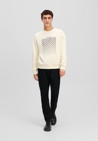 Crème sweatshirt met een gestructureerd zwart grafisch ontwerp op de voorkant, gecombineerd met zwarte broeken en zwarte laarzen. Eenvoudige en moderne silhouet.