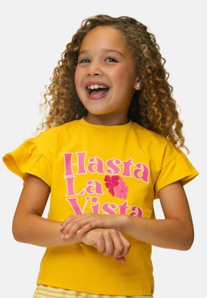 Glunderend meisje met krullend haar, gekleed in een geel shirt met de tekst "Hasta La Vista", staand met de handen gevouwen voor zich tegen een witte achtergrond.