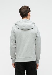 Jeune personne portant un sweat à capuche gris clair et un pantalon noir, vue de dos sur un fond uni.