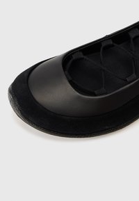 Crna baletna baletanka sa zaobljenim vrhom, glatkim i antilop teksturama te elastičnim križnim trakama preko otvorenog dijela za stopalo.