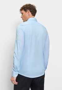 Man facing away wearing a light blue, long-sleeve, thin-striped dress shirt and black pants against a plain background.