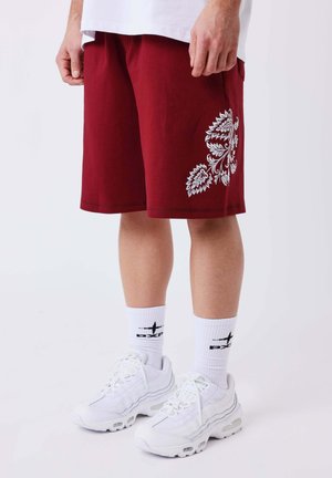 Persona con pantalones cortos rojos con estampado floral blanco, calcetines deportivos blancos y zapatillas blancas, de pie sobre un fondo claro y liso.