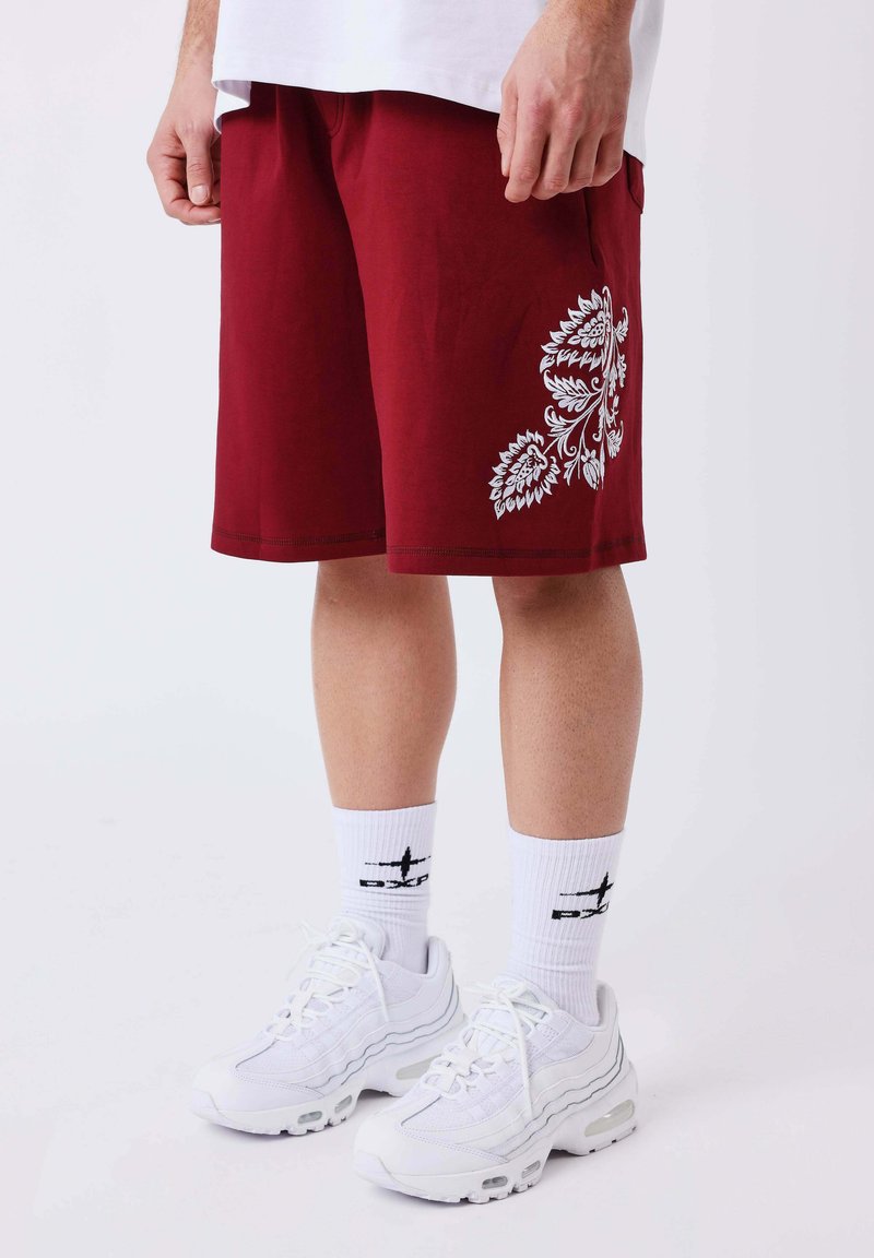 Personne portant un short rouge avec un imprimé floral blanc, des chaussettes de sport blanches et des baskets blanches, debout sur un fond clair uni.