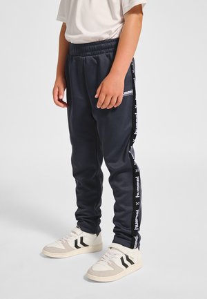 Granatowe spodnie dresowe z elastycznym pasem, ozdobione białymi logo po bokach i zwężanymi ściągaczami. Para beżowo-czarnych sneakersów.