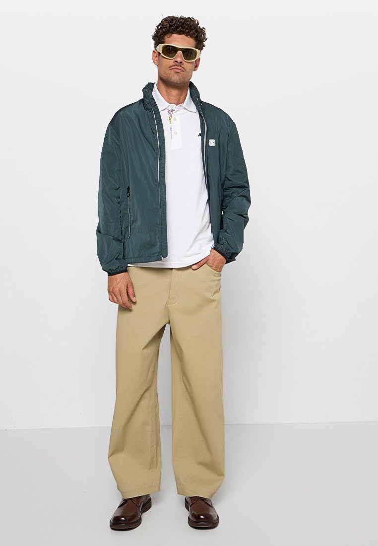 Uomo che indossa una giacca verde con zip, una camicia polo bianca, pantaloni beige a gamba larga, scarpe marroni e occhiali da sole con lenti gialle, in piedi contro uno sfondo neutro.