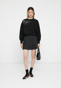 Svart genser med blomsterbroderi, parret med en grå plissert skirt. Modellen holder en liten svart håndveske, iført svarte sokker og to-farget sko.