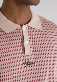 Beige knitted polo shirt with a textured pattern of small red shapes, featuring a beige collar and two visible buttons. "Prohibited" is embroidered in black.