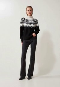 Svart-hvitt strikket genser med geometrisk mønster, høy krage og lange ermer, parret med mørke vide jeans og svarte hæler.