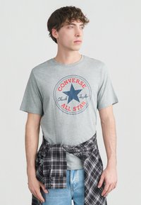 Νέος άνδρας που φοράει γκρι μπλουζάκι Converse All Star και ανοιχτόχρωμο μπλε τζιν, με ένα μαύρο και άσπρο καρό πουκάμισο δεμένο γύρω από τη μέση του.