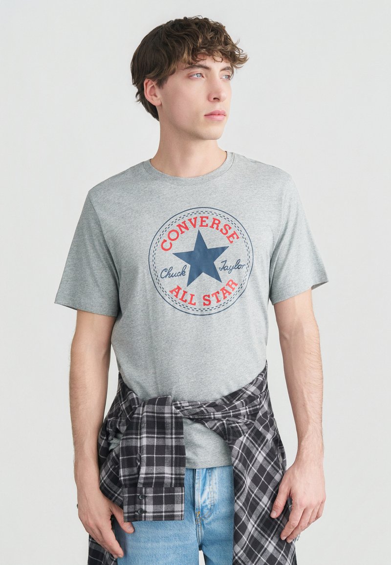 Νέος άνδρας που φοράει γκρι μπλουζάκι Converse All Star και ανοιχτόχρωμο μπλε τζιν, με ένα μαύρο και άσπρο καρό πουκάμισο δεμένο γύρω από τη μέση του.