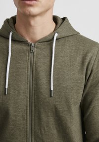 Olivengrønn zip-up hettegenser med hvite snorer, med en jevn tekstur og en metallglidelås. Nærbilde som viser kragen og hette-detaljene.