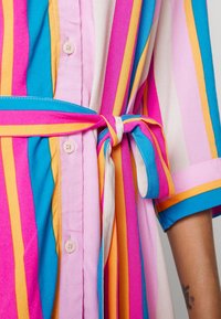 Chemise rayée présentant des couleurs bleue, rose, orange et blanche, avec une taille nouée, des boutons décoratifs et des manches courtes. Texture de tissu lisse.