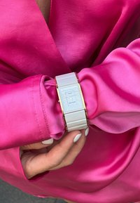 Reloj rectangular plateado con esfera blanca y detalles en oro, llevado en una muñeca cubierta por una manga de satén rosa brillante. Correa texturizada.