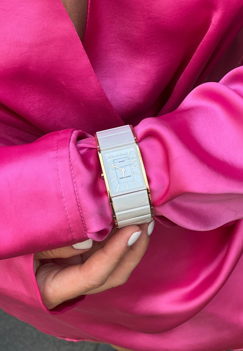 Reloj rectangular plateado con esfera blanca y detalles en oro, llevado en una muñeca cubierta por una manga de satén rosa brillante. Correa texturizada.
