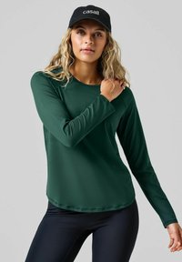 Zielony sportowy top z długim rękawem, wykonany z gładkiego materiału, z okrągłym dekoltem i przeszytymi brzegami. Połączony z czarnymi legginsami.