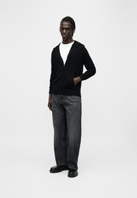 Sweat à capuche noir zippé avec poches avant, porté sur un t-shirt blanc. Associé à un jean gris en denim et des chaussures noires. Design simple et minimaliste.