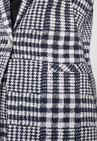 Tkanina w wzór houndstooth w kolorze granatowym i białym charakteryzuje się teksturowaną powierzchnią oraz dopasowanym krojem, z kieszonką na piersi i srebrnym guzikiem.
