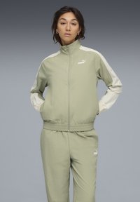 Lichtgroen trainingspak van gladde stof, met een hoge kraag, ritssluiting en elastische manchetten. Witte accenten op de mouwen en het logo.