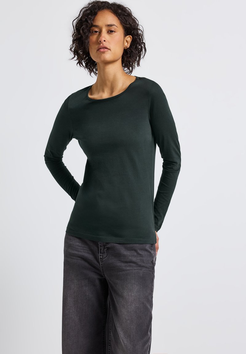 Donna con capelli ricci che indossa un top a maniche lunghe verde scuro e jeans neri sbiaditi, in piedi con le mani dietro la schiena contro uno sfondo neutro.