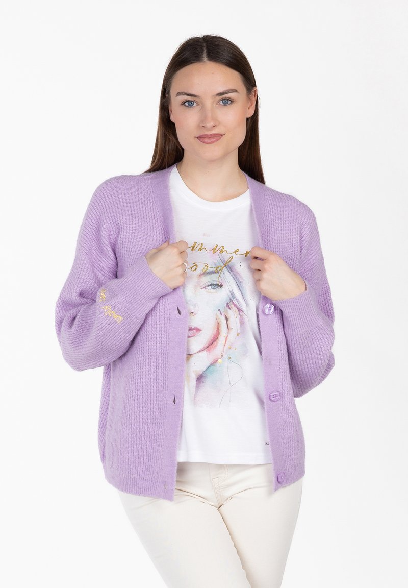 Frau trägt eine fliederfarbene Strickjacke über einem weißen Hemd mit Aquarell-Gesichtsprint und beigefarbenen Hosen, steht vor einem weißen Hintergrund.