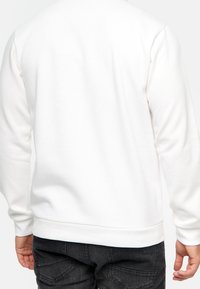 Hvid sweatshirt med lange ærmer, fast tekstur, ribbede manchetter og kant. Båret med mørke jeans, viser ryggen.