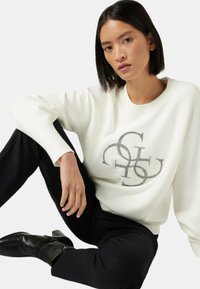 Sudadera blanca con diseño de logo de rhinestones negros, cuello redondo, puños de canalé, combinada con pantalones negros y botas de tobillo. Textura suave.