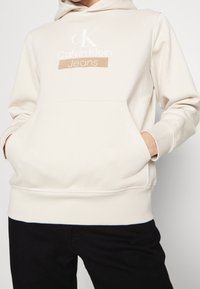 Beige huvtröja med kenguruficka, prydd med vit Calvin Klein-logotyp och textilstruktur. Ribbad mudd och nederkant.