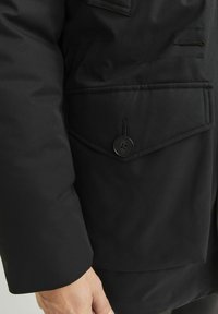 Abrigo negro con una textura suave, con un gran botón y un bolsillo de parche en la parte frontal. Detalle con cremallera y tejido duradero.