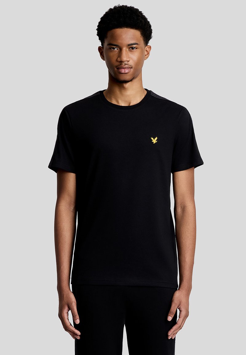 Junger Mann trägt ein schlichtes schwarzes T-Shirt mit einem kleinen gelben Vogel-Logo auf der linken Brust, steht vor einem einfarbigen grauen Hintergrund.