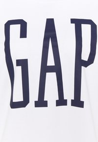 T-shirt en coton blanc affichant un grand logo « GAP » bleu marine en lettres capitales et épaisses sur le devant. Texture lisse, col rond.