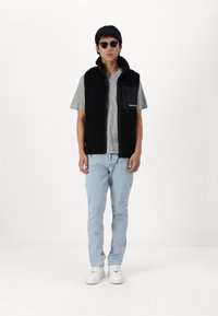 Zwarte fleece vest met een hoge kraag, rits aan de voorkant en een zak op de linkerborst. Gecombineerd met een grijs T-shirt en lichtblauwe jeans. Witte sneakers.