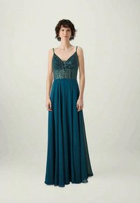 Moteris, apsirengusi grindimis siekiančia teal spalvos suknele su blizgučių korpusu ir plonomis petnešėlėmis, stovi prieš paprastą šviesią fono.