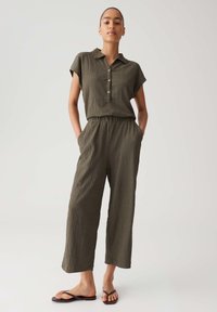 Olive green, textured jumpsuit with a collared neck, short sleeves, button closure, and elastic waist; paired with flat sandals.