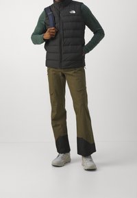 Man i svart ärmlös puffväst, grön långärmad tröja, olivgröna cargo-byxor med svarta ärmslut, ljusa vandringsskor och en ryggsäck.